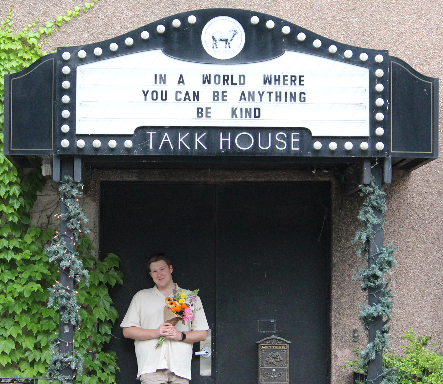 Hunter Hodge under a sign that says 'In a world where you can be anything, be kind.'.
